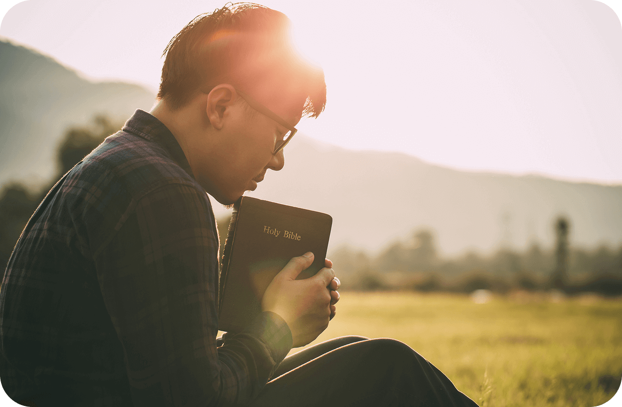 A person sits outdoors on a grassy field, holding a book titled "Holy Bible" close, with sunlight and hills in the background.