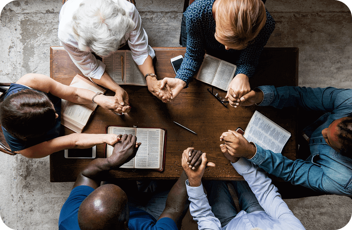People on their personal faith journeys sit around a wooden table, holding hands and reading books, as seen from above.