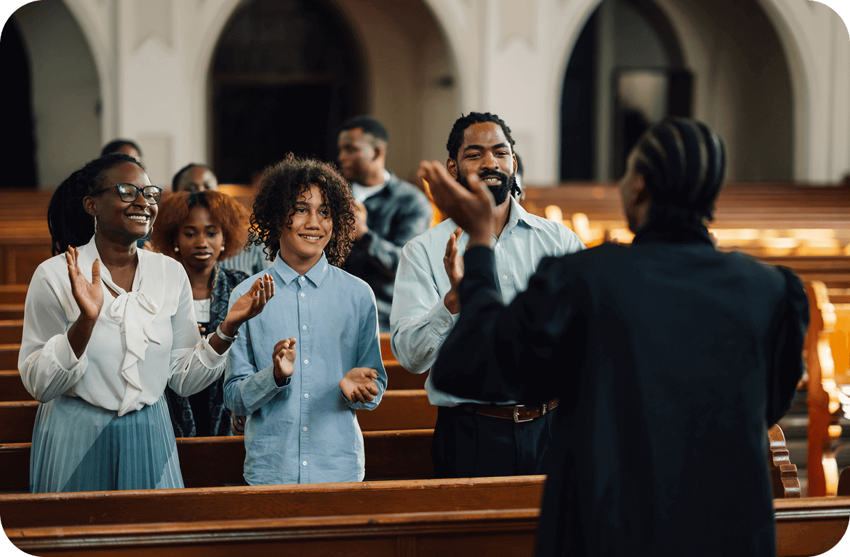 A group of people stands in a church, clapping and smiling as they listen to a speaker share stories of personal faith journeys. The atmosphere is warm and welcoming, with wooden pews and arches adding to the serene backdrop.