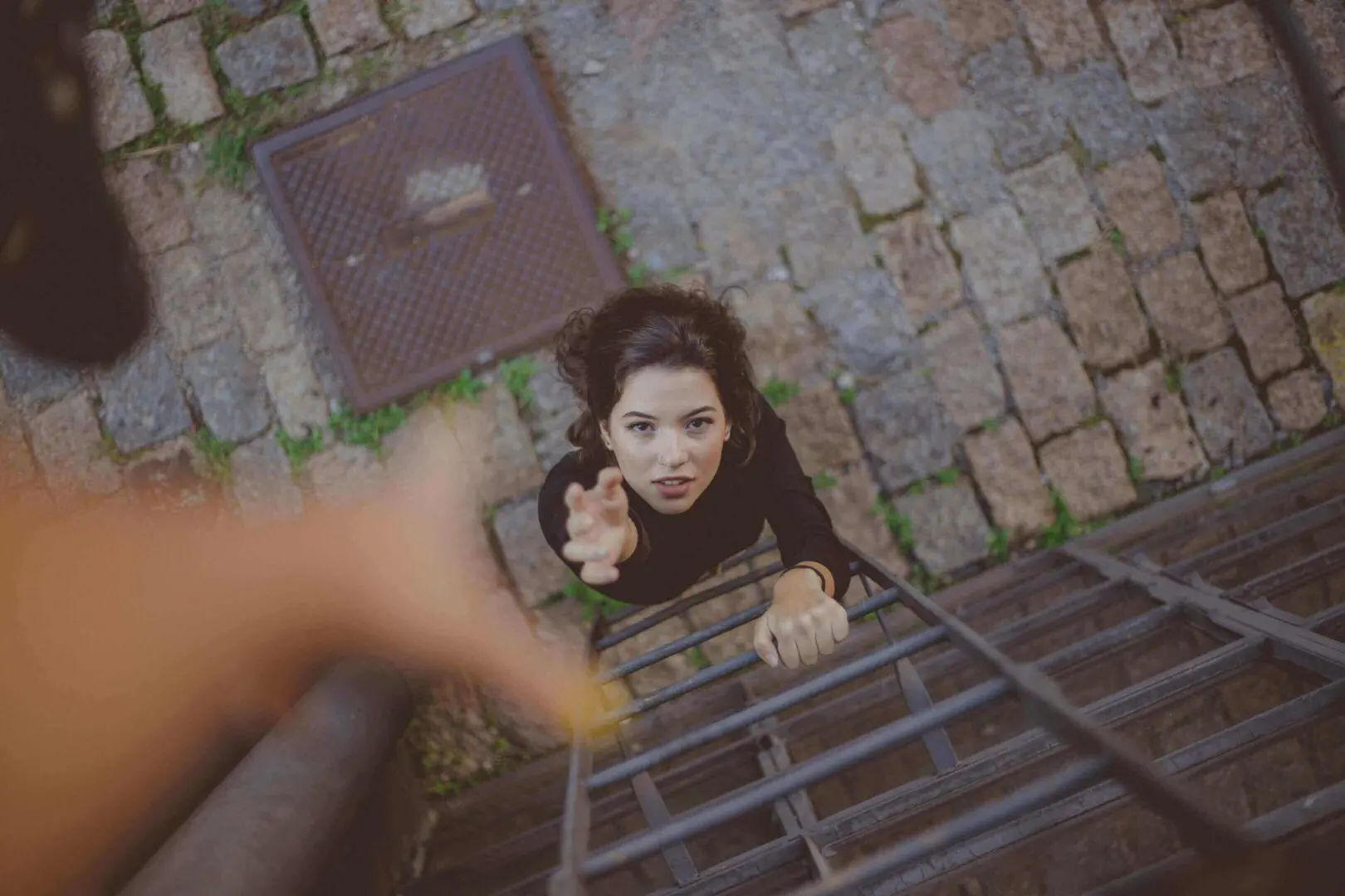 A woman reaches up a metal ladder with a hand extended toward her.