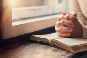 Person praying with hands clasped over an open book near a window.