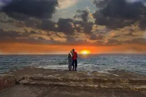 Couple embracing by the ocean at sunset with dramatic clouds.
