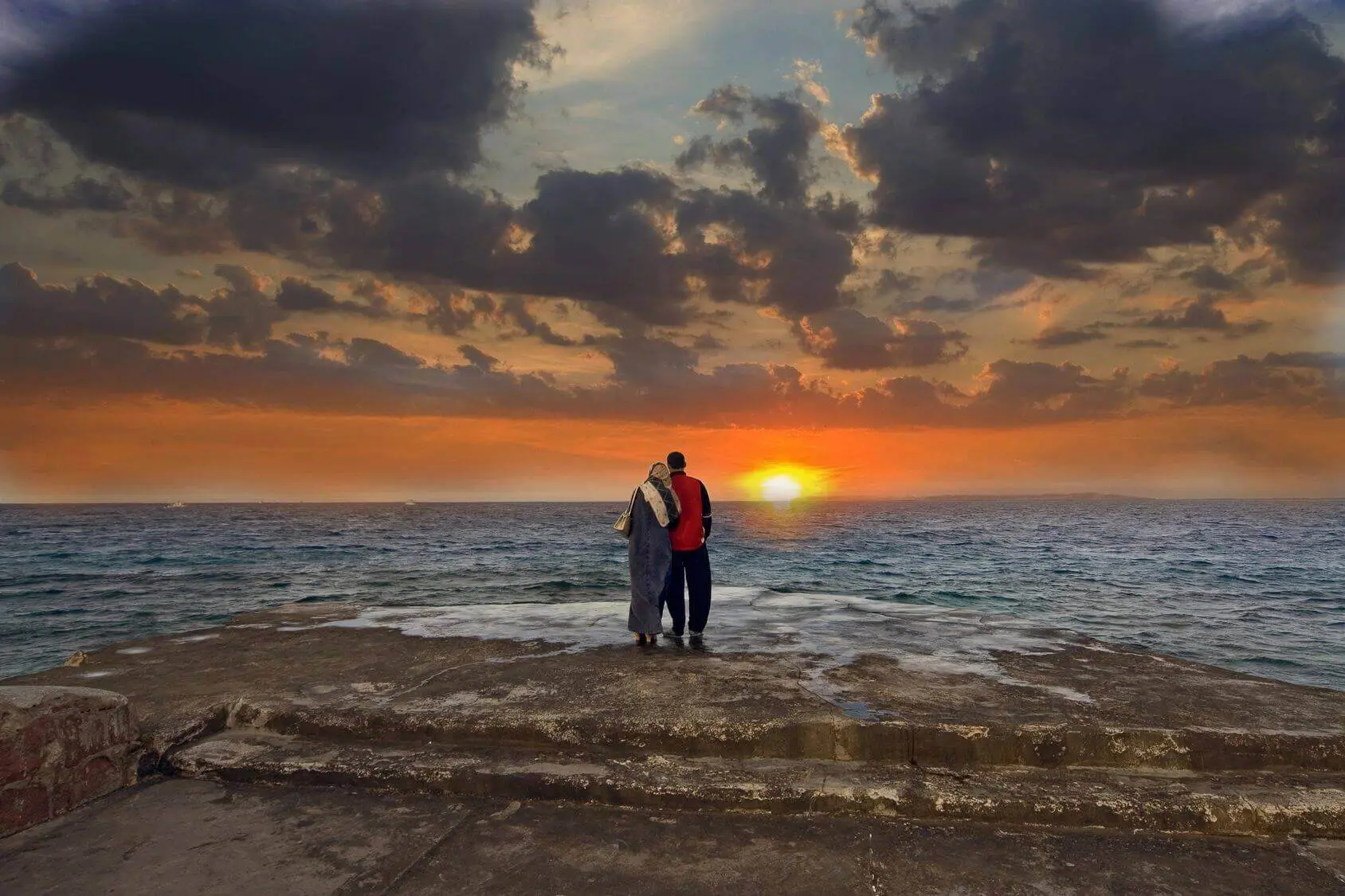 Couple embracing by the ocean at sunset with dramatic clouds.