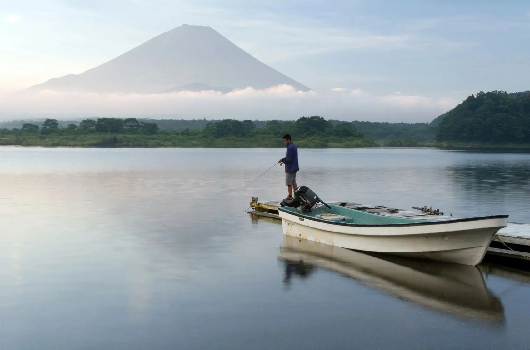 A person fishing from a boat on calm water near a mountain.