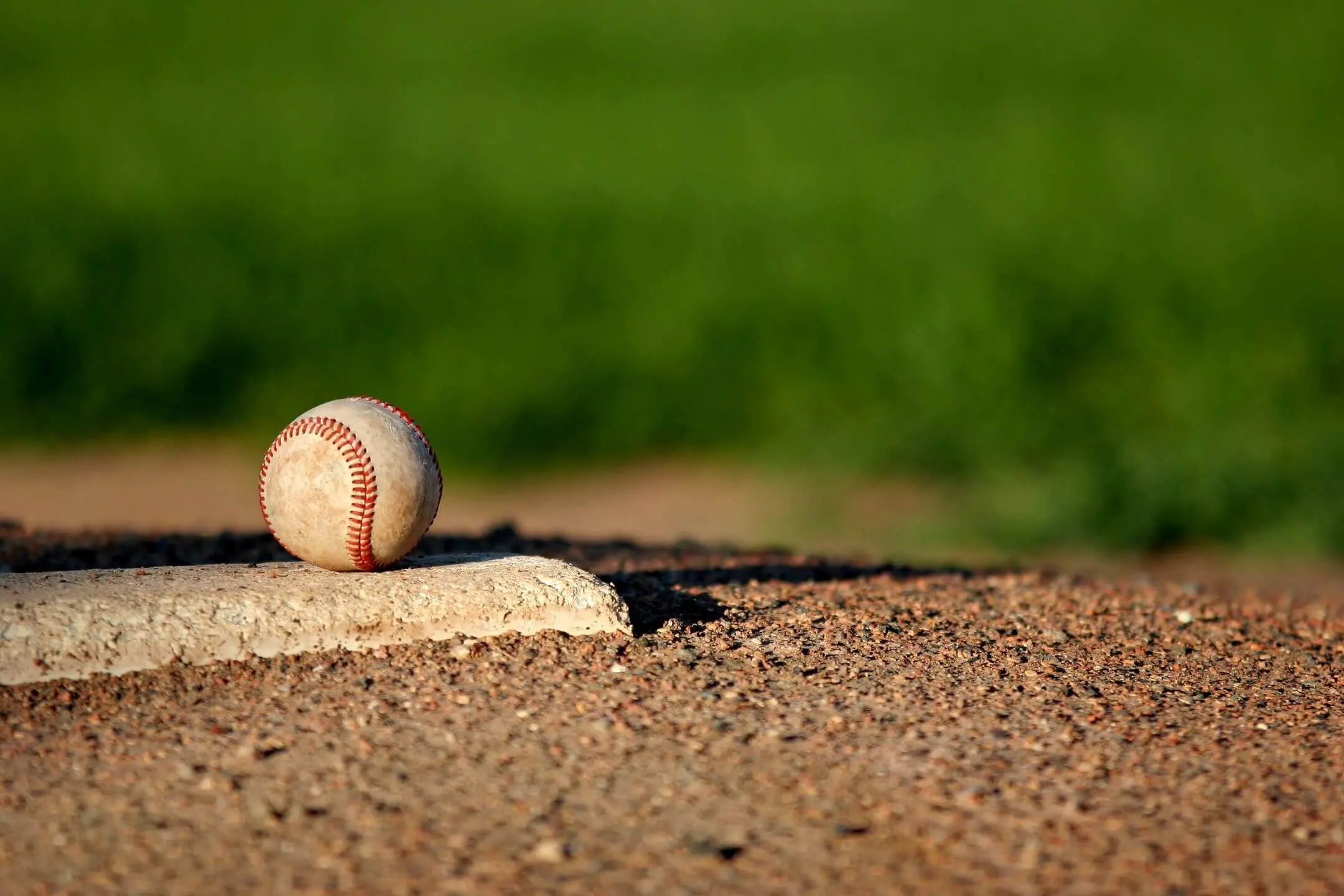 A baseball resting on home plate on a dirt field with green grass in the background.