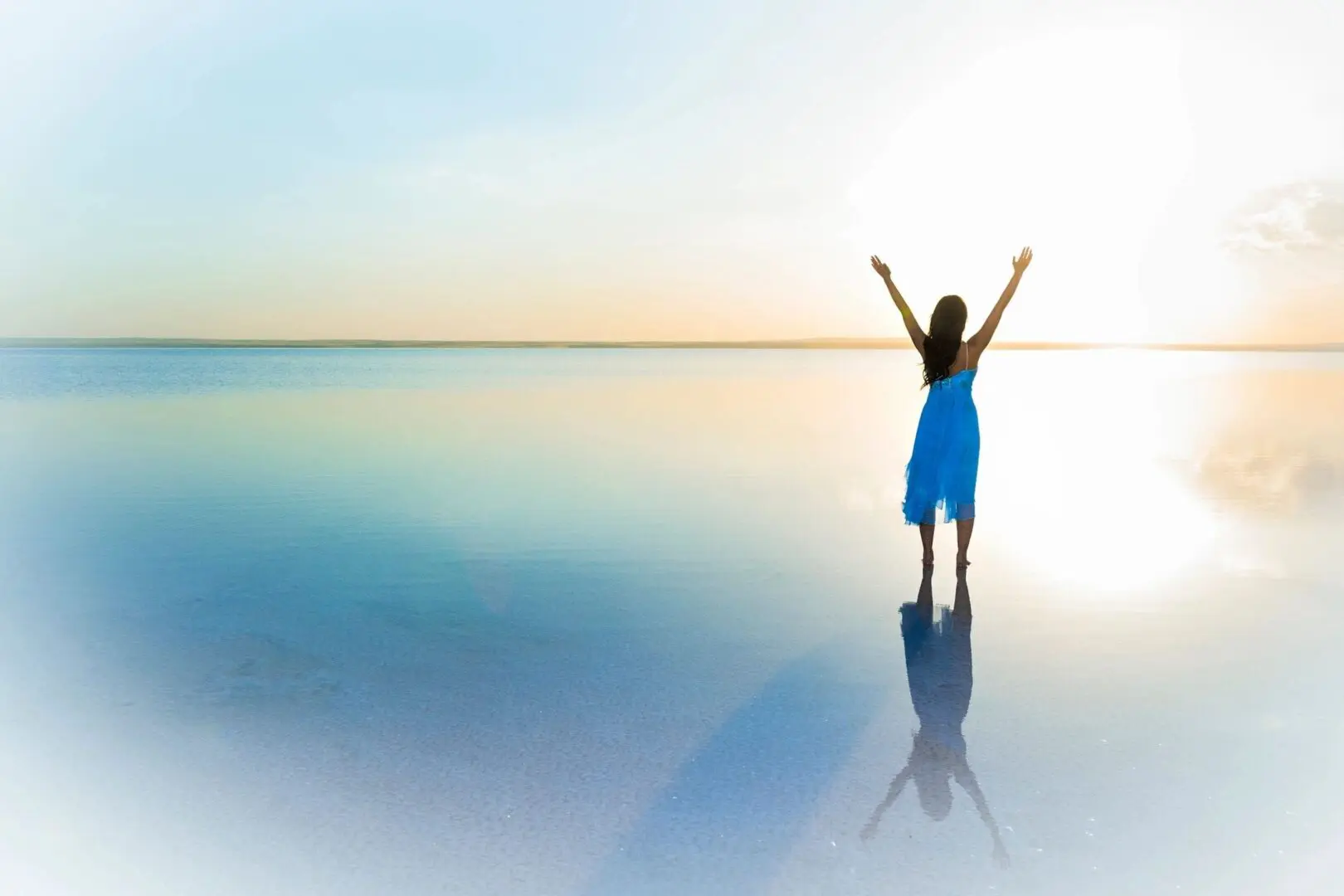 A person in a blue dress stands with arms raised on a reflective water surface at sunrise.