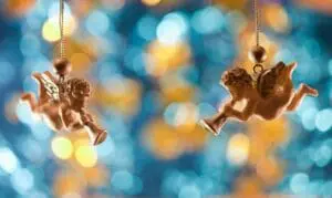 Two cherubic angel ornaments hanging against blurred colorful lights.