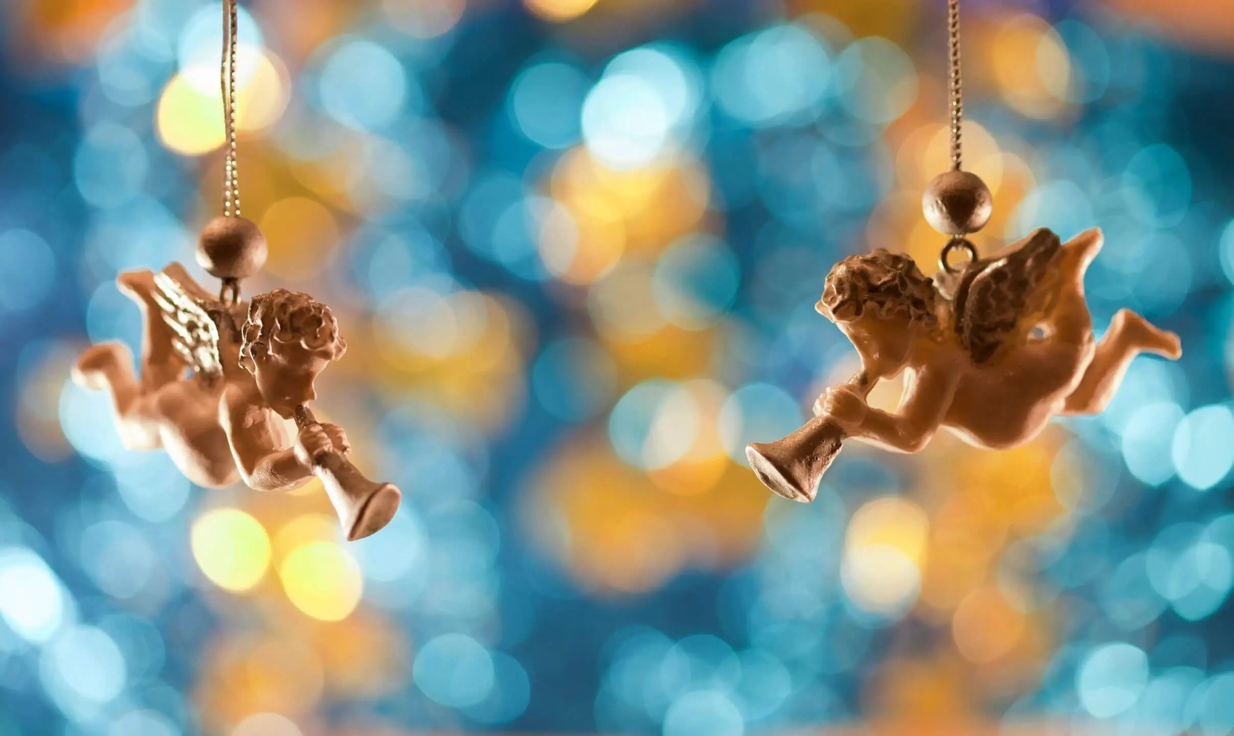 Two cherubic angel ornaments hanging against blurred colorful lights.