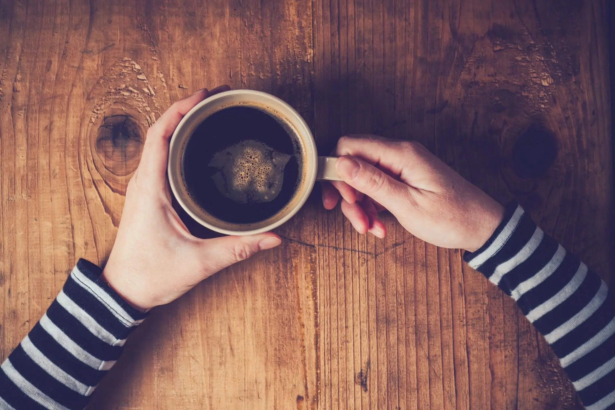 Hands holding a cup of coffee over a wooden table.