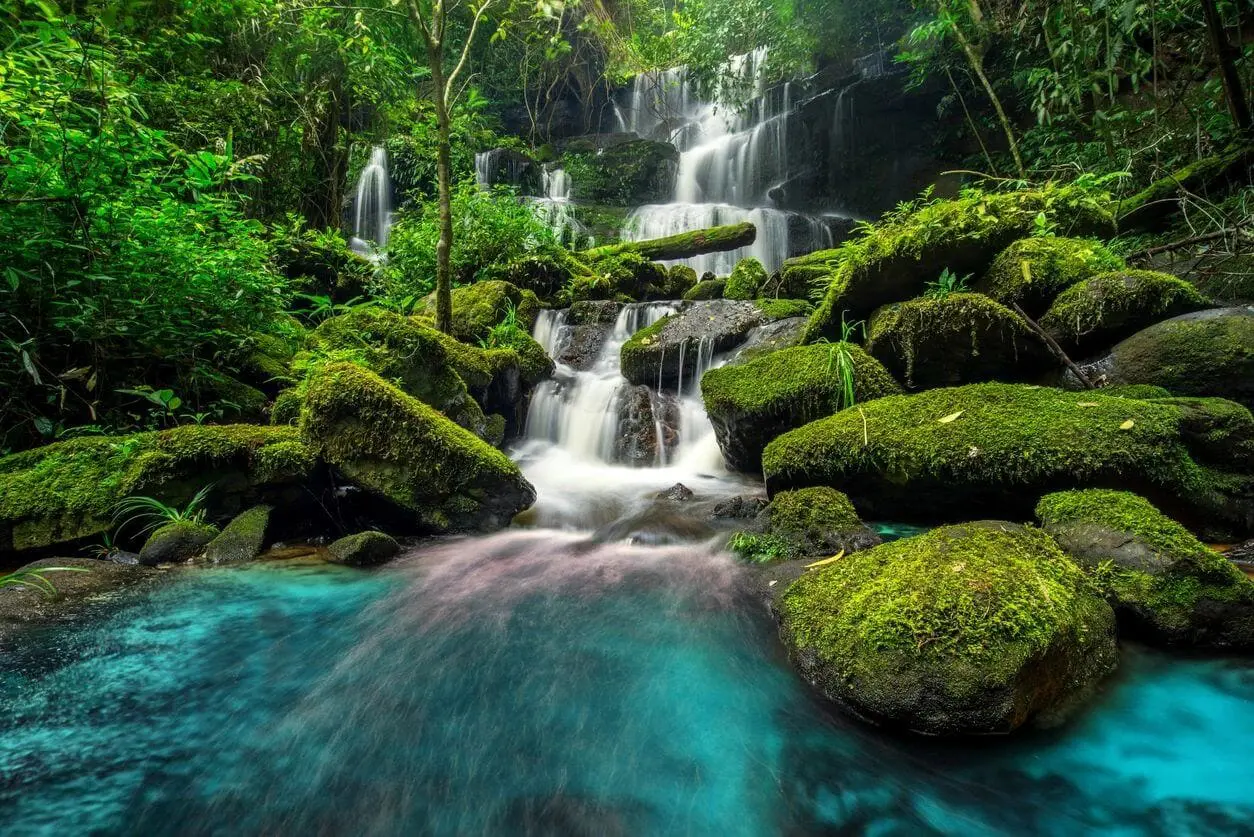 A serene waterfall flows over moss-covered rocks into clear blue water.