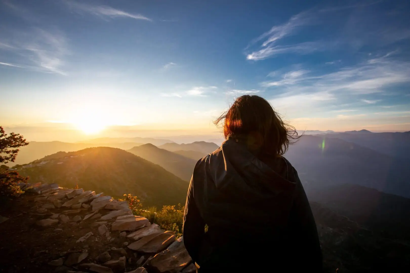 Person watching sunset over mountains from a rocky viewpoint.