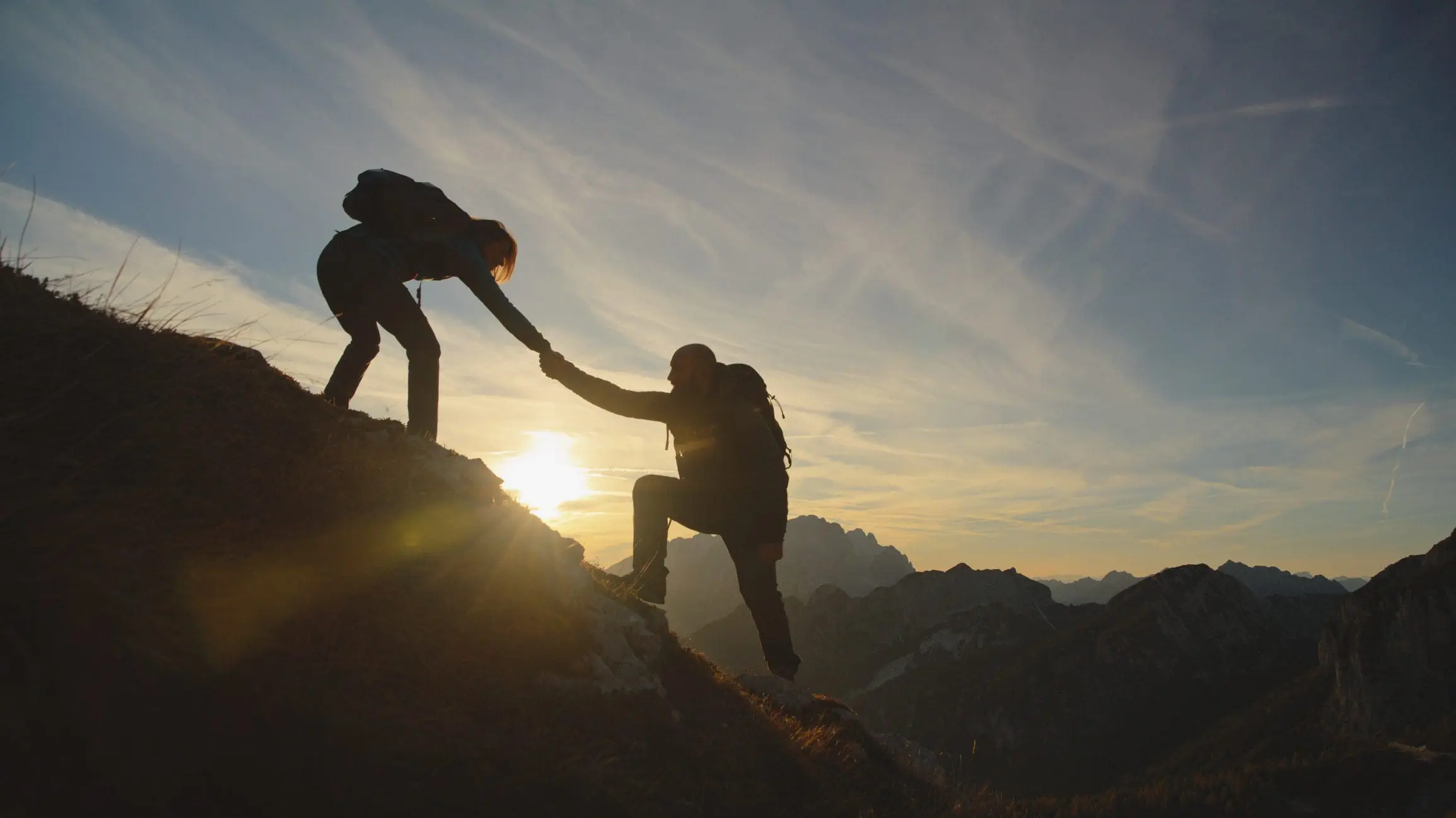 Two hikers helping each other climb a mountain at sunset.