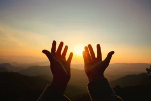 Hands reaching toward the sunset over mountains.