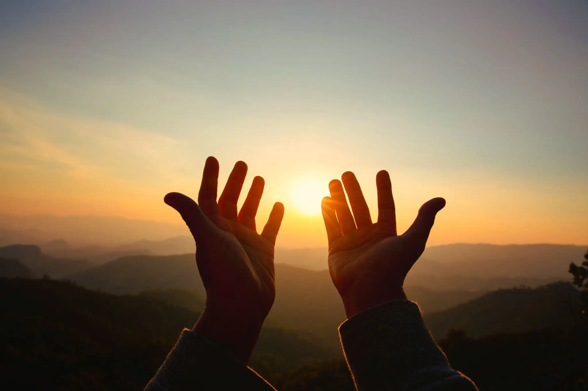 Hands reaching toward the sunset over mountains.