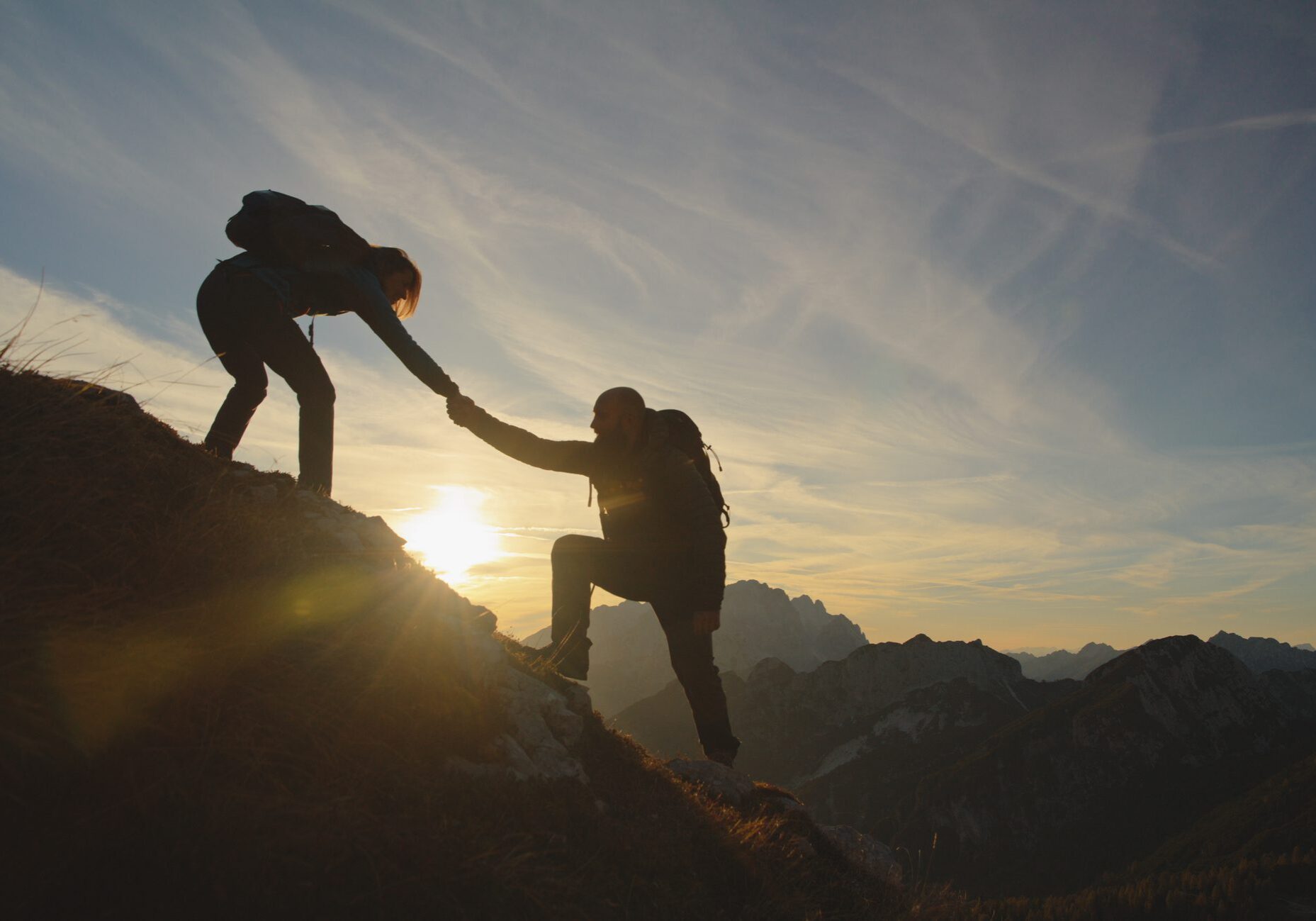 Two hikers helping each other climb a mountain at sunset.