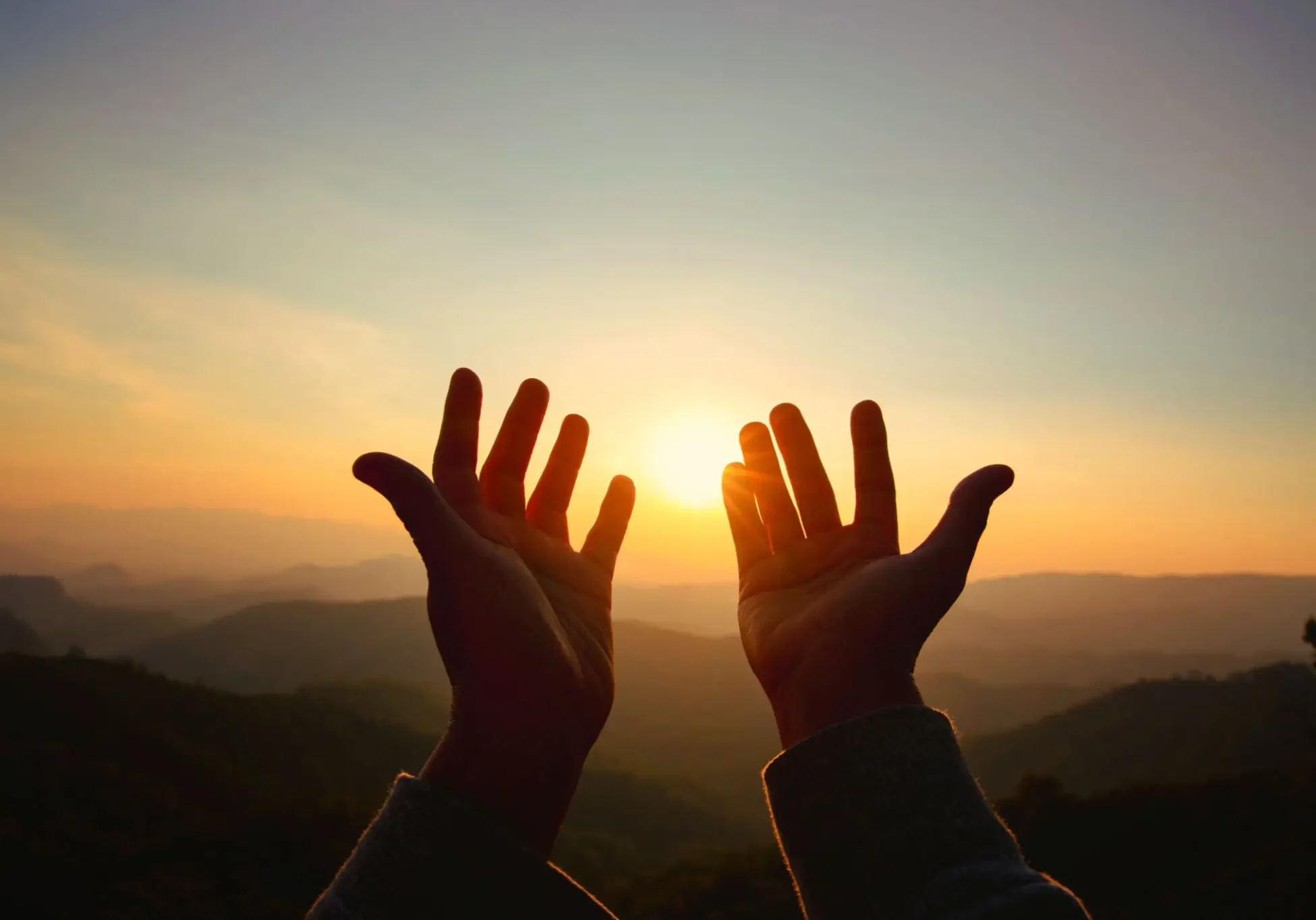 Hands reaching toward the sunset over mountains.