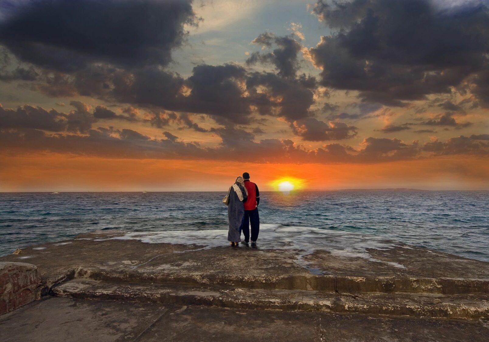 Couple embracing by the ocean at sunset with dramatic clouds.