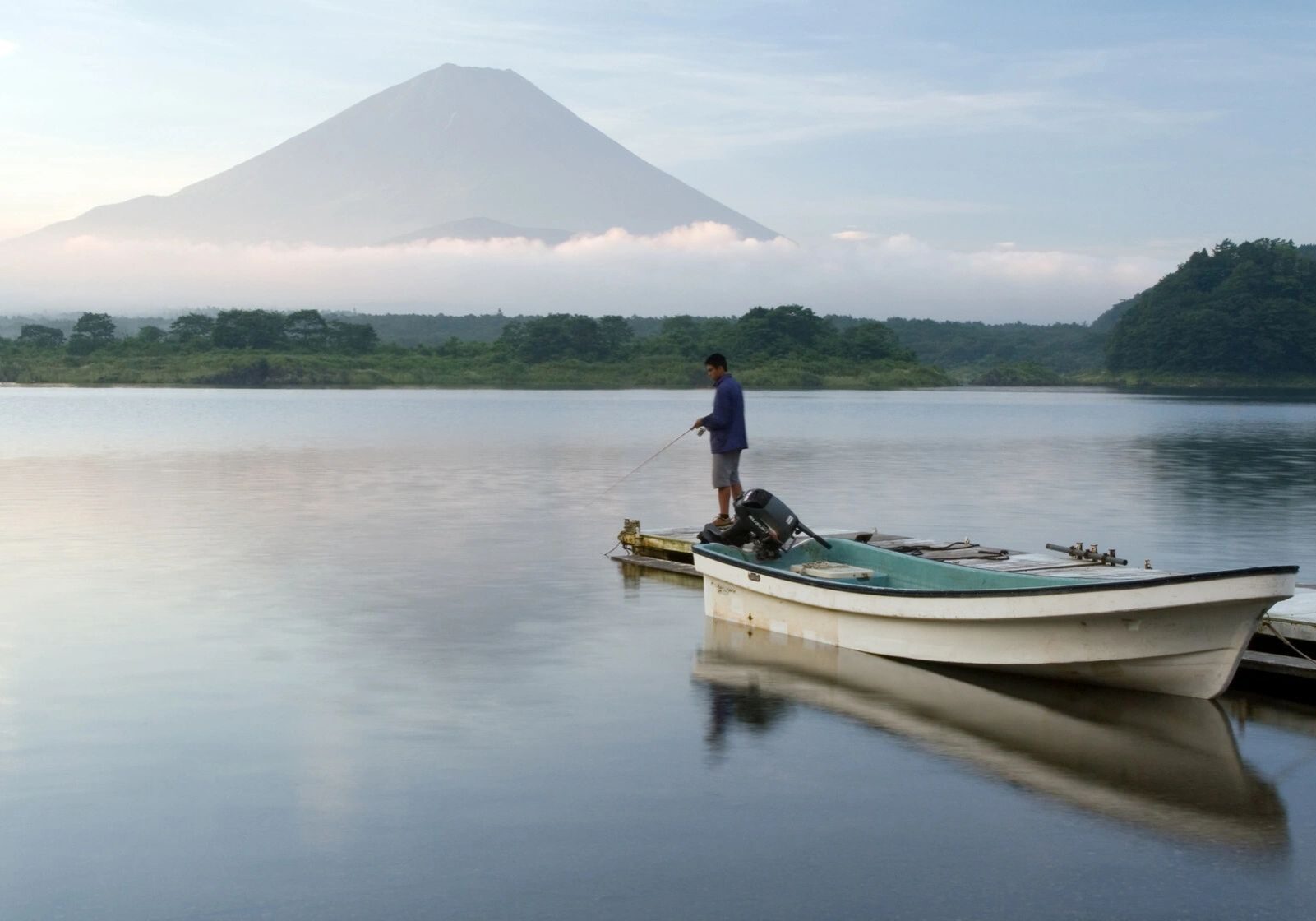 A person fishing from a boat on calm water near a mountain.