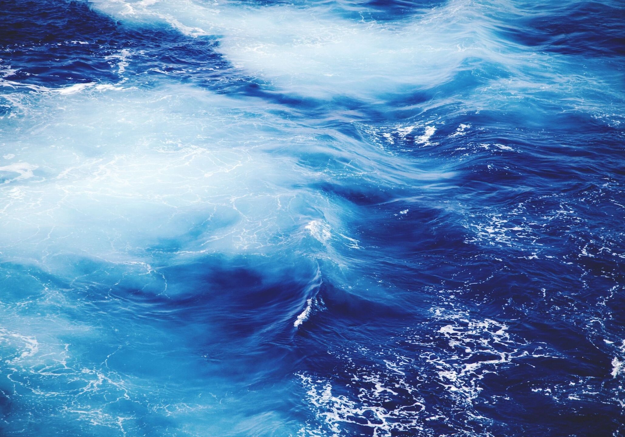 Swirling blue ocean waves with white foam under sunlight.