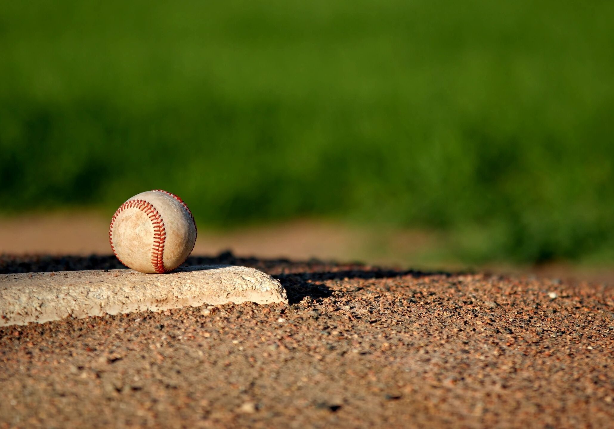 A baseball resting on home plate on a dirt field with green grass in the background.