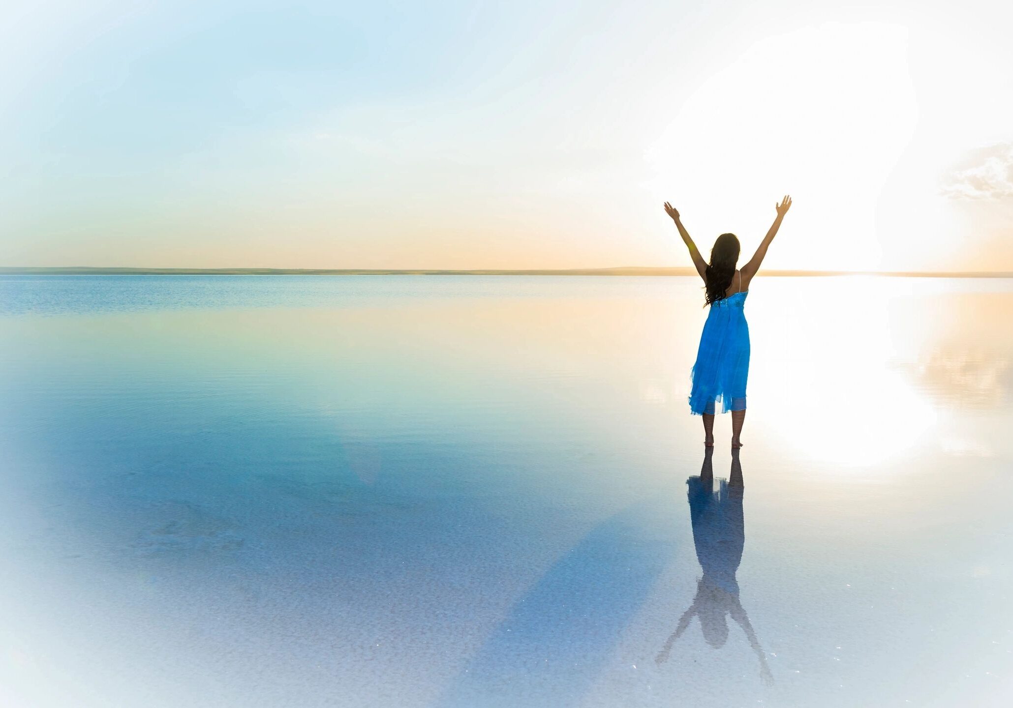 A person in a blue dress stands with arms raised on a reflective water surface at sunrise.