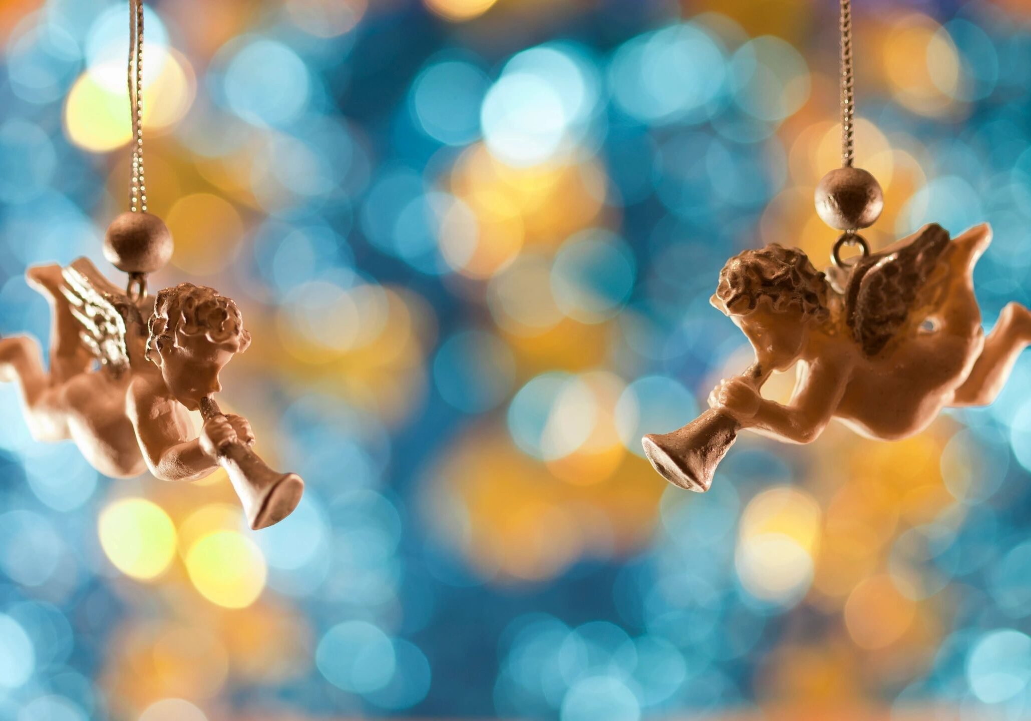 Two cherubic angel ornaments hanging against blurred colorful lights.