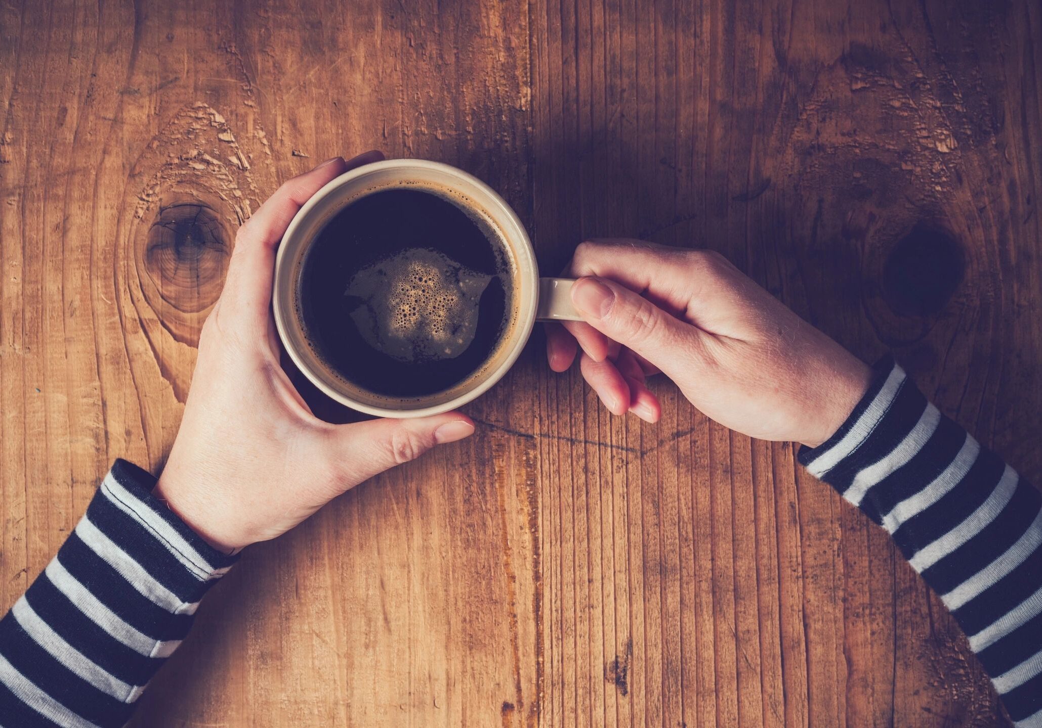 Hands holding a cup of coffee over a wooden table.