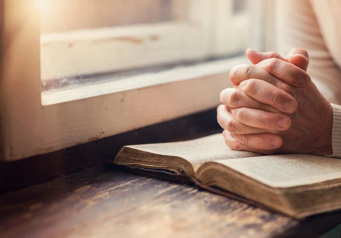 Person praying with hands clasped over an open book near a window.